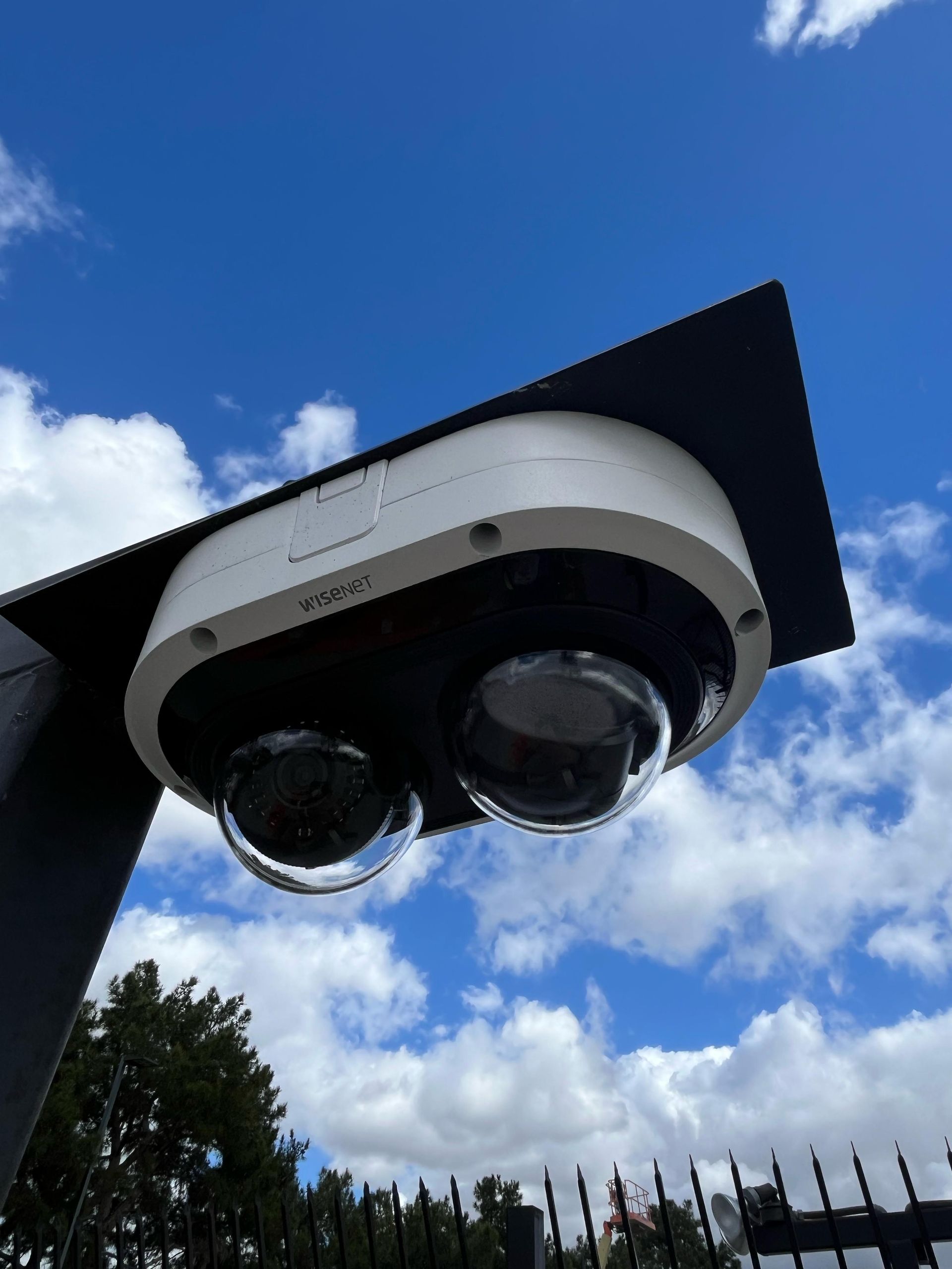 Dual security cameras on a black bracket against a blue sky with clouds.