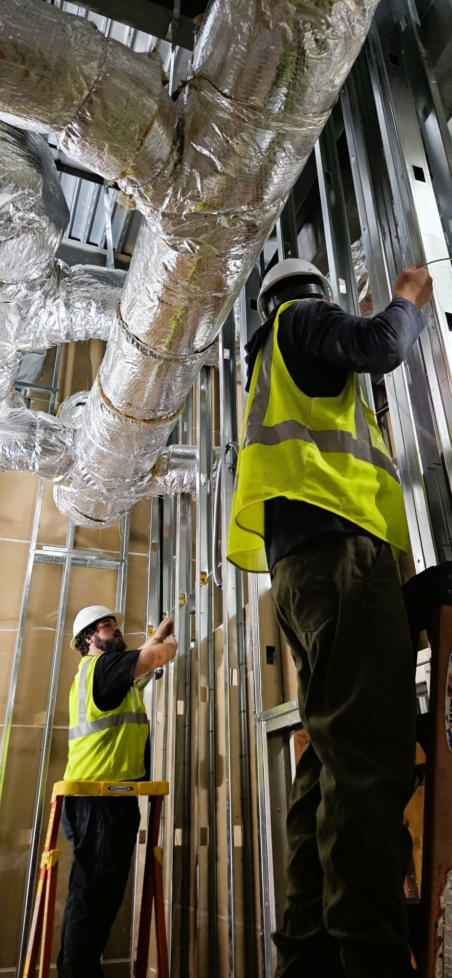 Construction workers installing wall frames near insulated pipes.