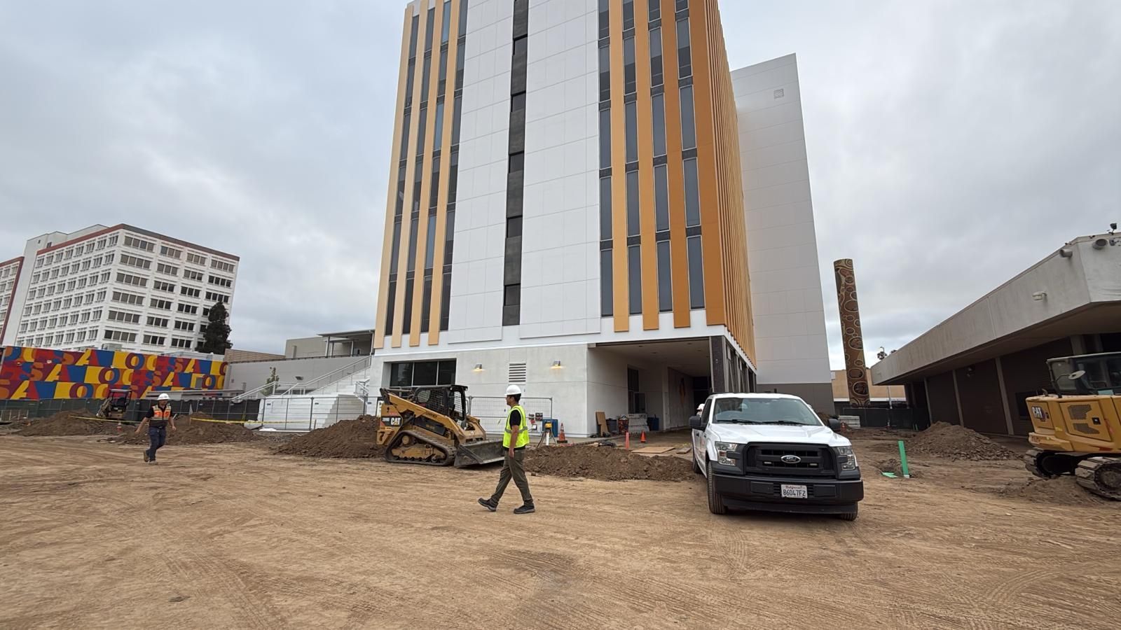 Construction site with a tall modern building and a worker walking. Brown dirt in foreground, cloudy sky.