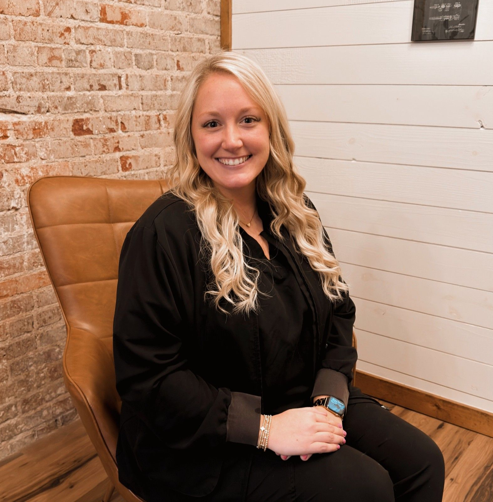 Woman in gray scrubs smiles, seated in a brown chair, brick and wood background.