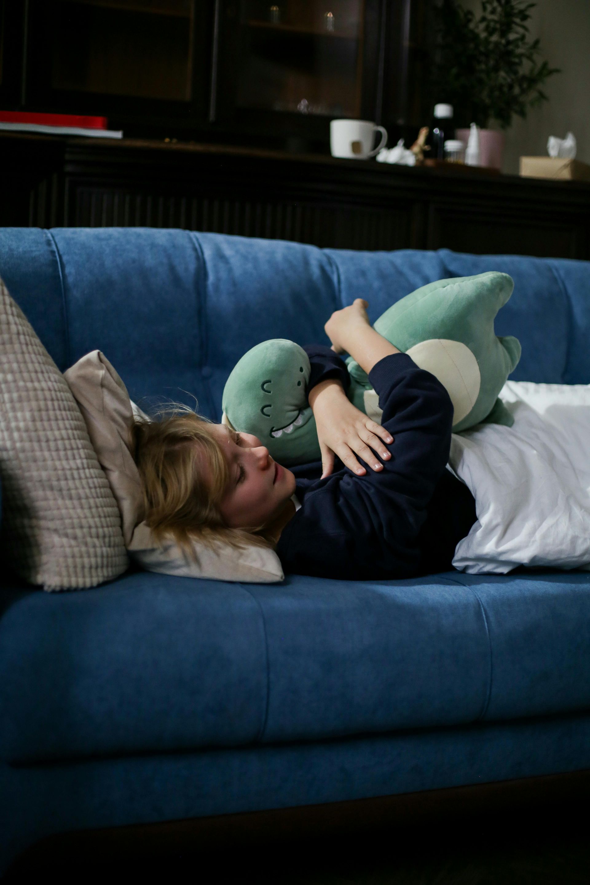 A woman is laying on a blue couch holding a stuffed dinosaur.