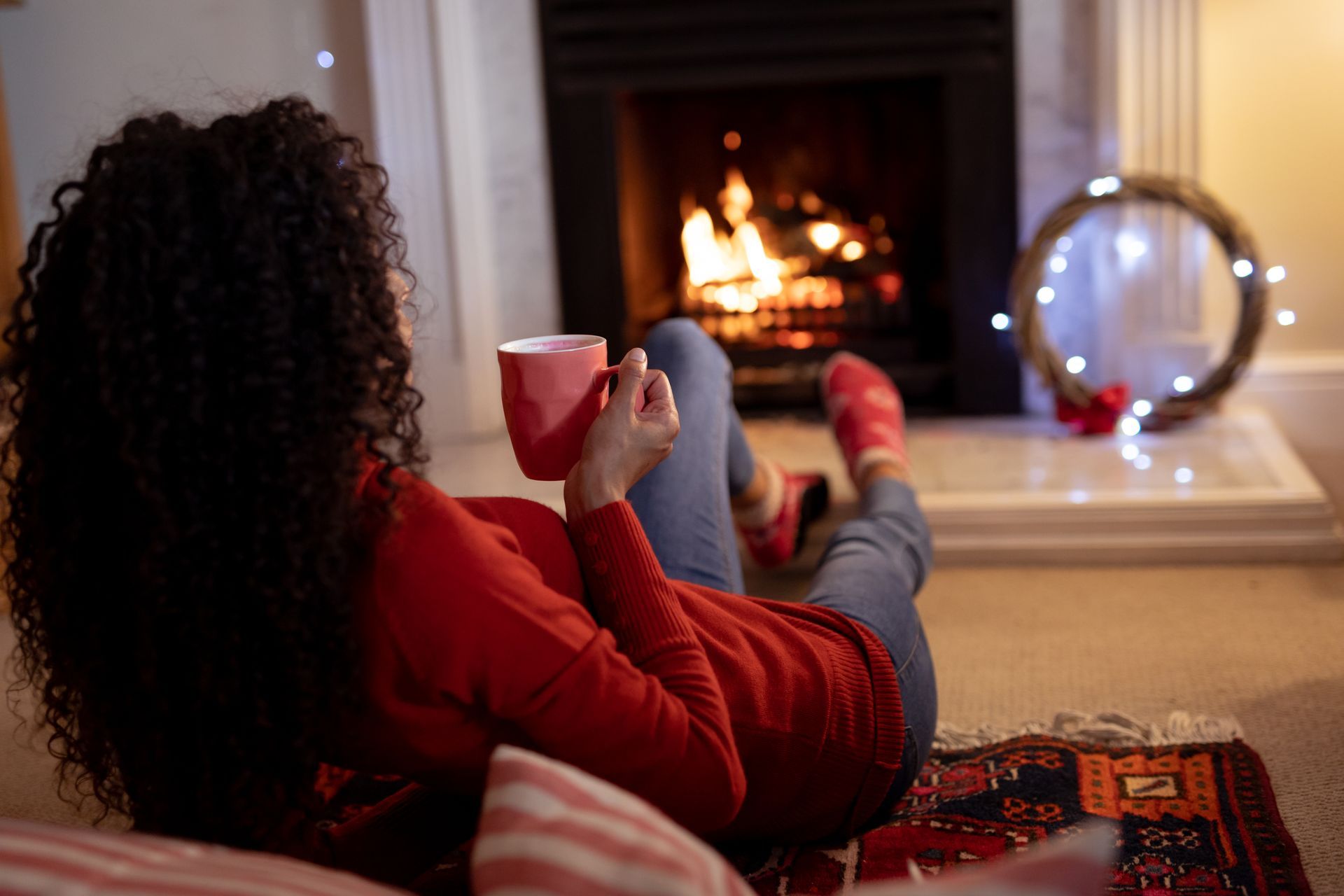 A woman is sitting on the floor drinking coffee in front of a fireplace.