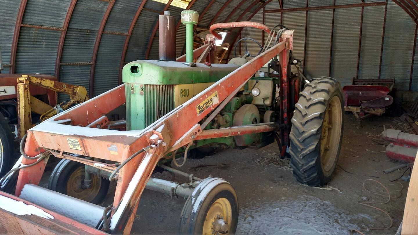 Old green tractor with front loader in a barn, with a large rear wheel visible.