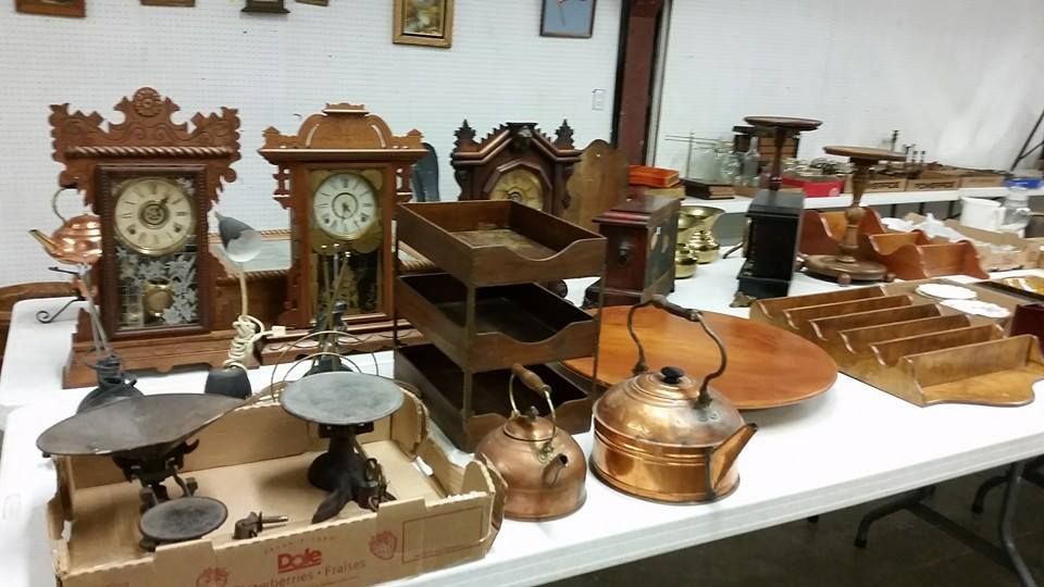 Table covered with assorted antique household items, including clocks, scales, and copper kettles in a room.