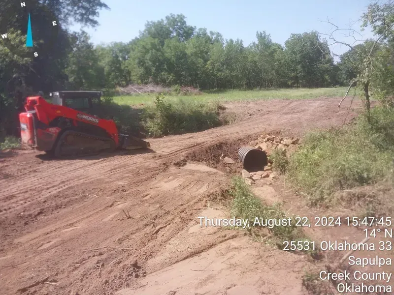 A bulldozer is driving down a dirt road next to a pipe.