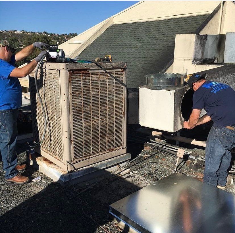Two men are working on an air conditioner on the roof of a building