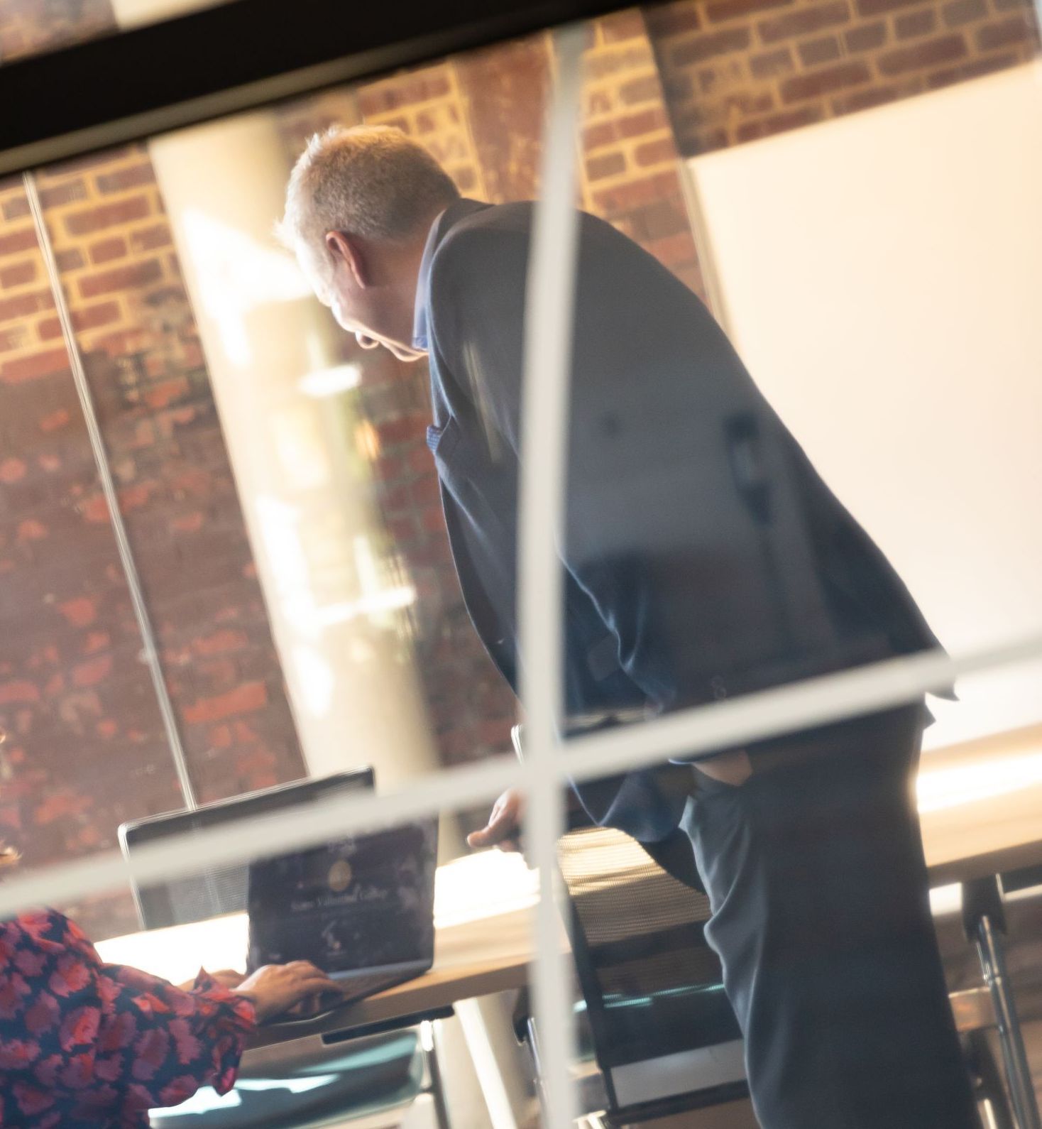 A man in a suit stands next to a woman using a laptop
