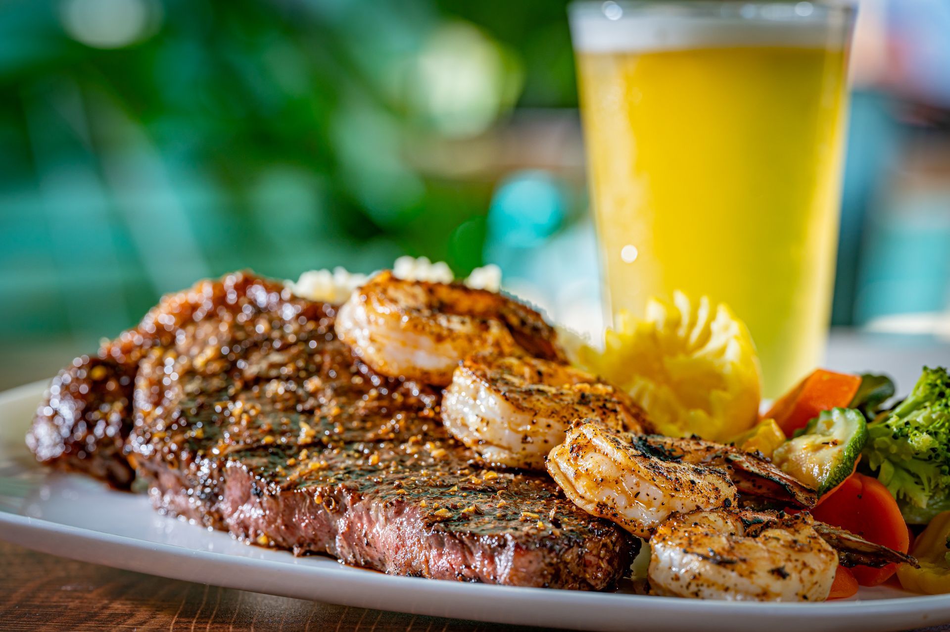 A plate of food with a steak , shrimp and vegetables next to a glass of beer.