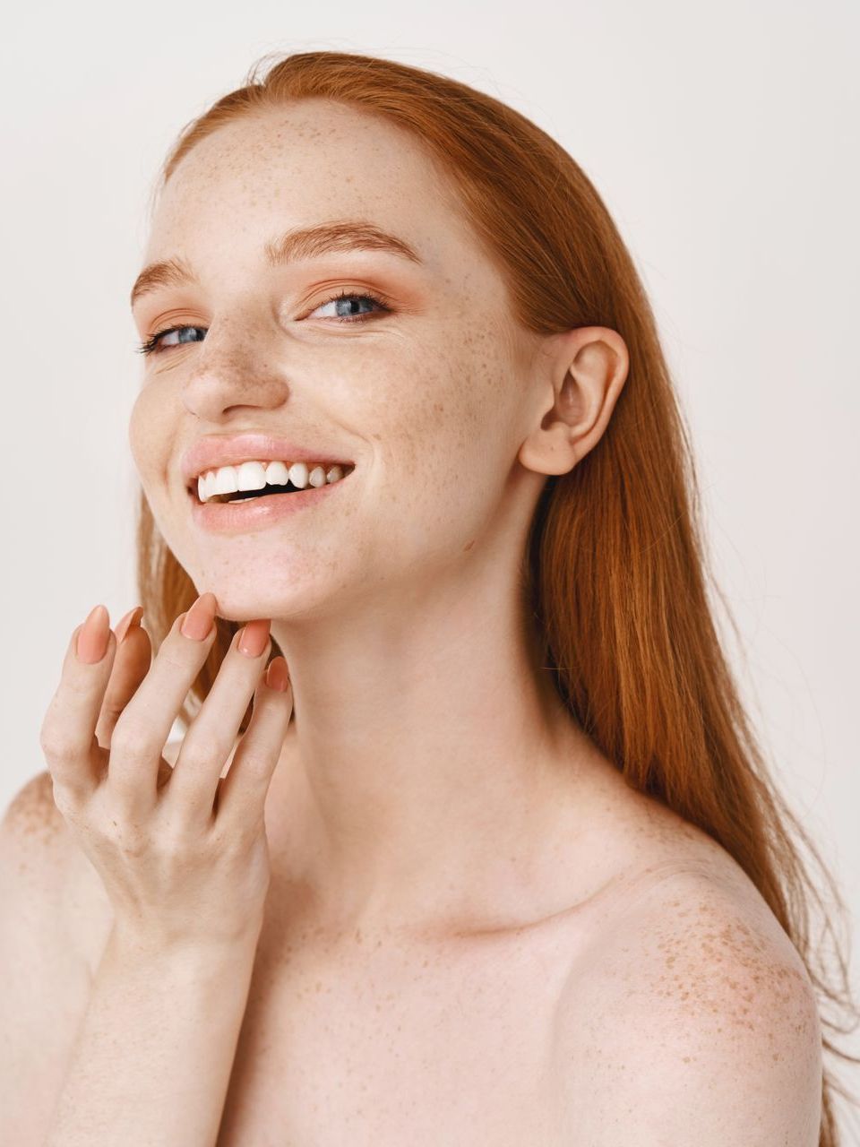 A woman with red hair and freckles is smiling and touching her face.