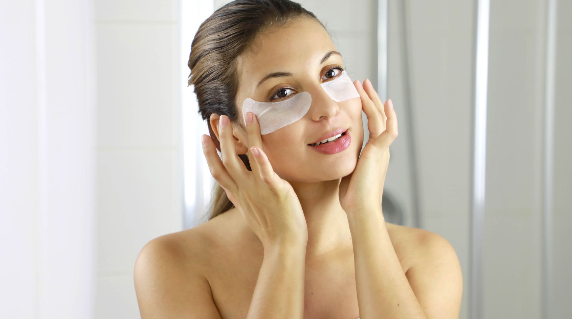 A woman is applying eye patches to her face in front of a mirror.