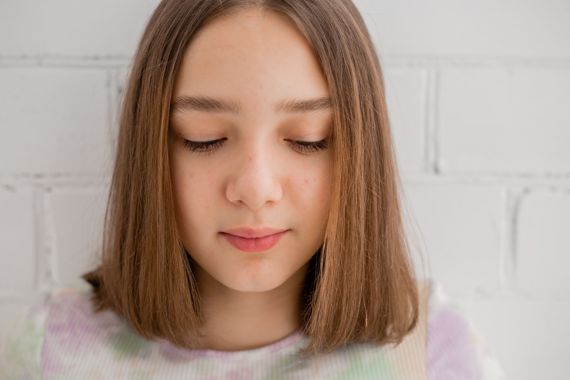 A young girl with her eyes closed is standing in front of a white brick wall.