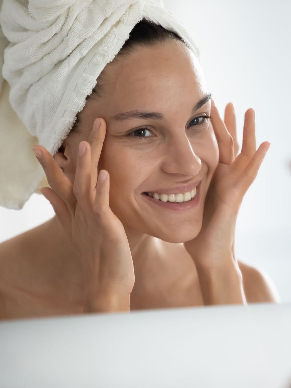 A woman with a towel wrapped around her head is looking at her face in the mirror.