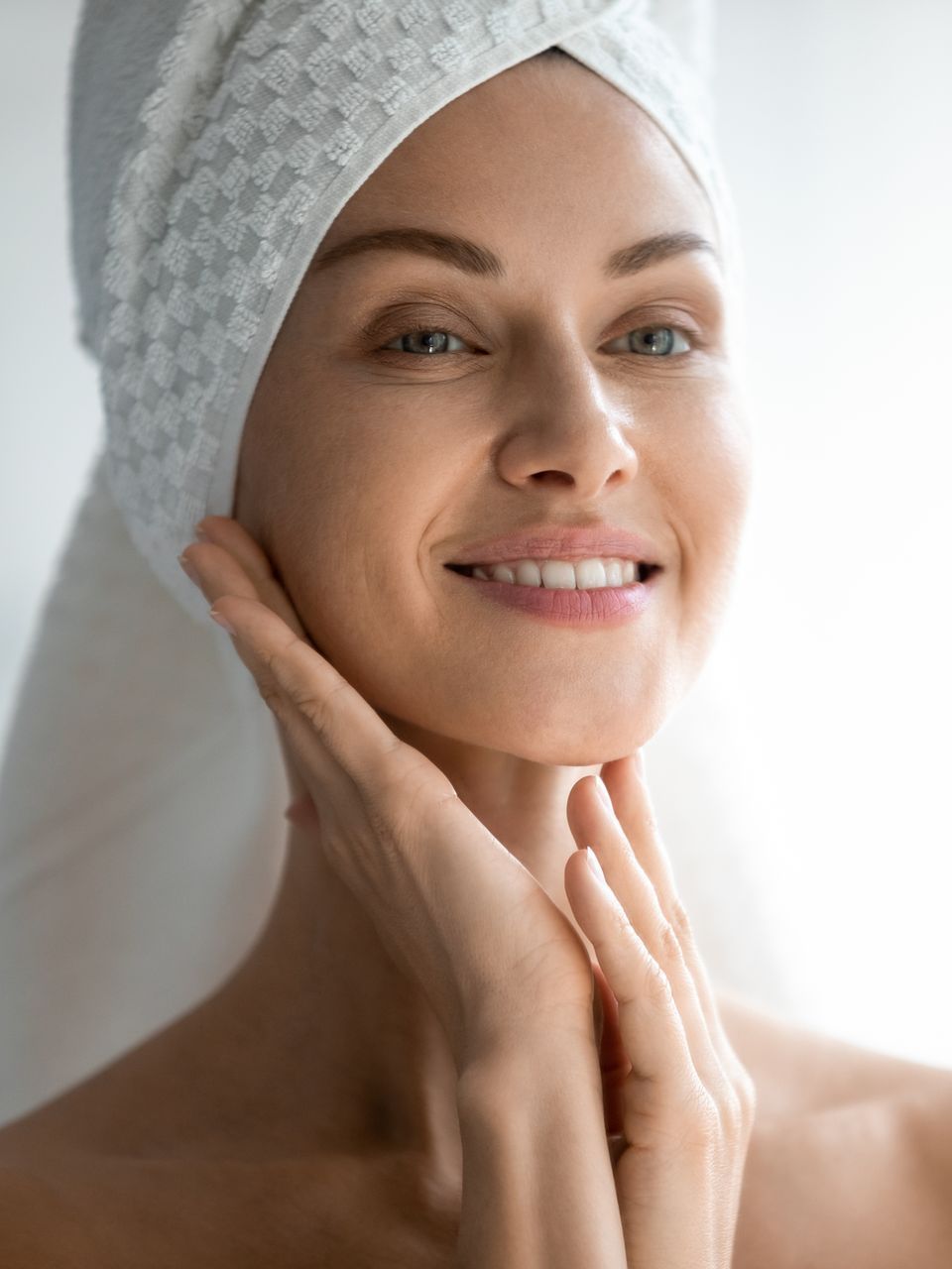 A woman with a towel wrapped around her head is smiling and touching her face.