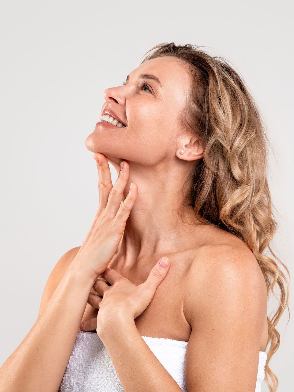 A woman wrapped in a towel is touching her neck and smiling.