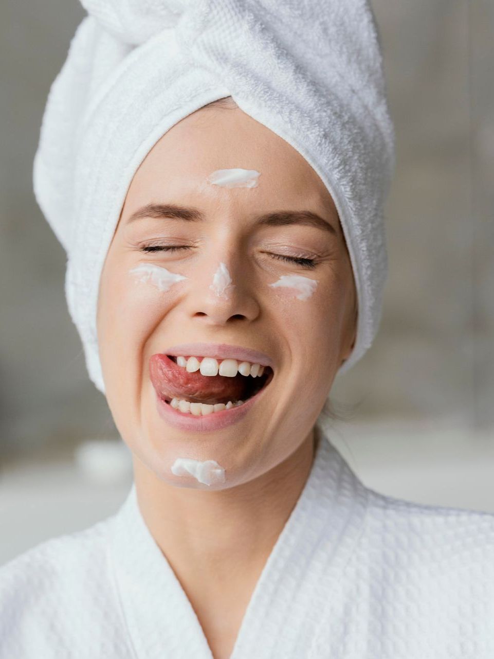 A woman with a towel wrapped around her head is laughing with cream on her face.