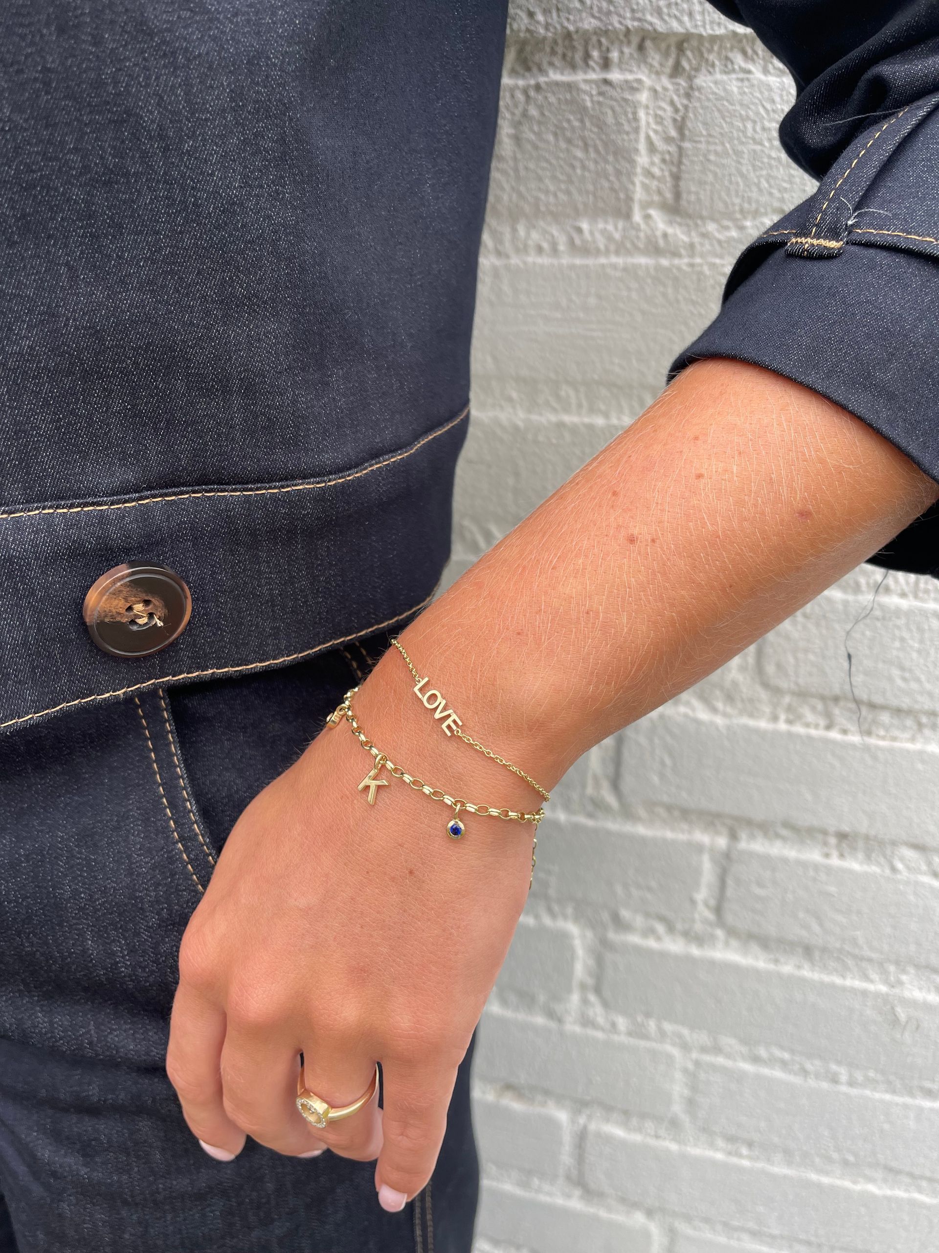 Een vrouw draagt ​​een gouden armband en een ring op haar arm, tegen een achtergrond van een donker spijkerjack en een witte bakstenen muur.