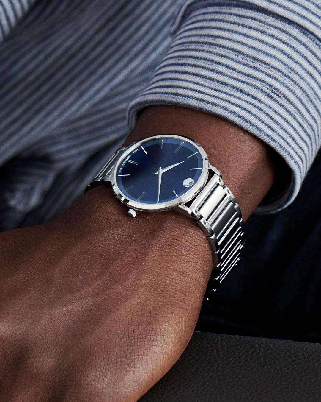 Zilveren horloge met een donkerblauwe wijzerplaat op iemands pols, tegen een gestreept shirt.