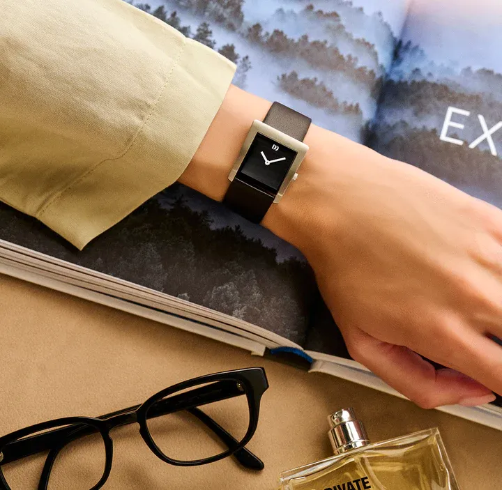 Persoon draagt ​​een zwart rechthoekig horloge om zijn pols. Ernaast een bril, een open boek en parfum.