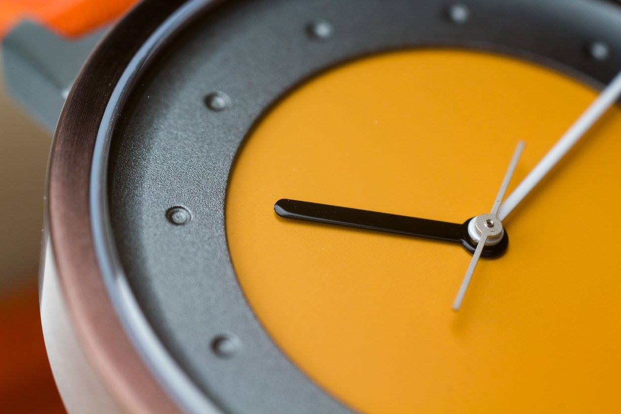 Close-up van een polshorloge met een oranje wijzerplaat en zwarte wijzers die ongeveer 3:10 aangeven.