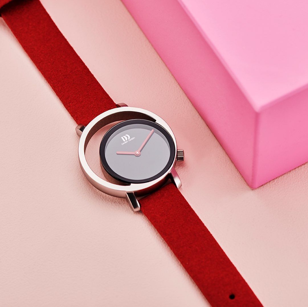 Horloge met rode band, grijze wijzerplaat en rode wijzers, op een roze achtergrond.