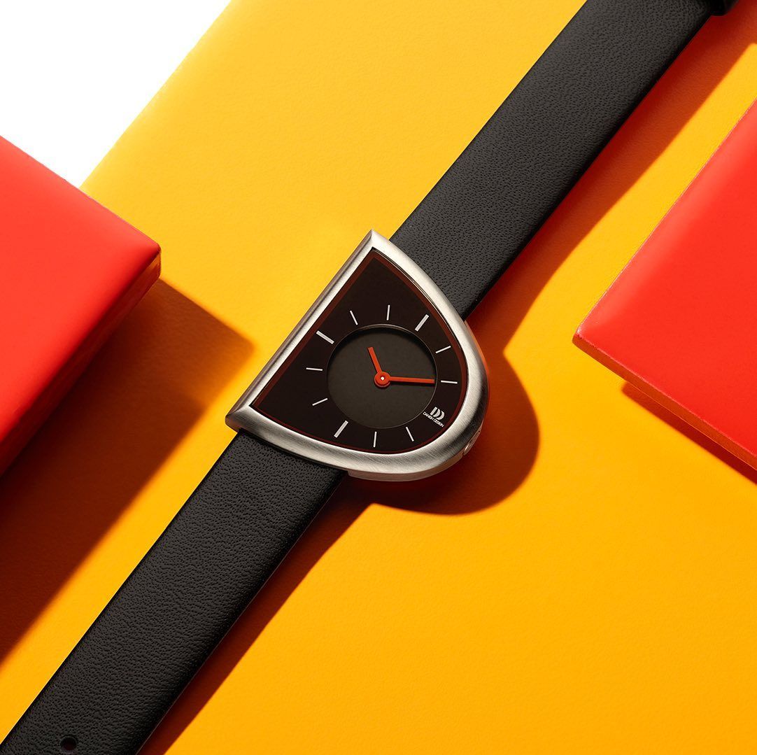 Zwart horloge met een unieke, halfronde wijzerplaat en zwarte band op een gele achtergrond met rode objecten.