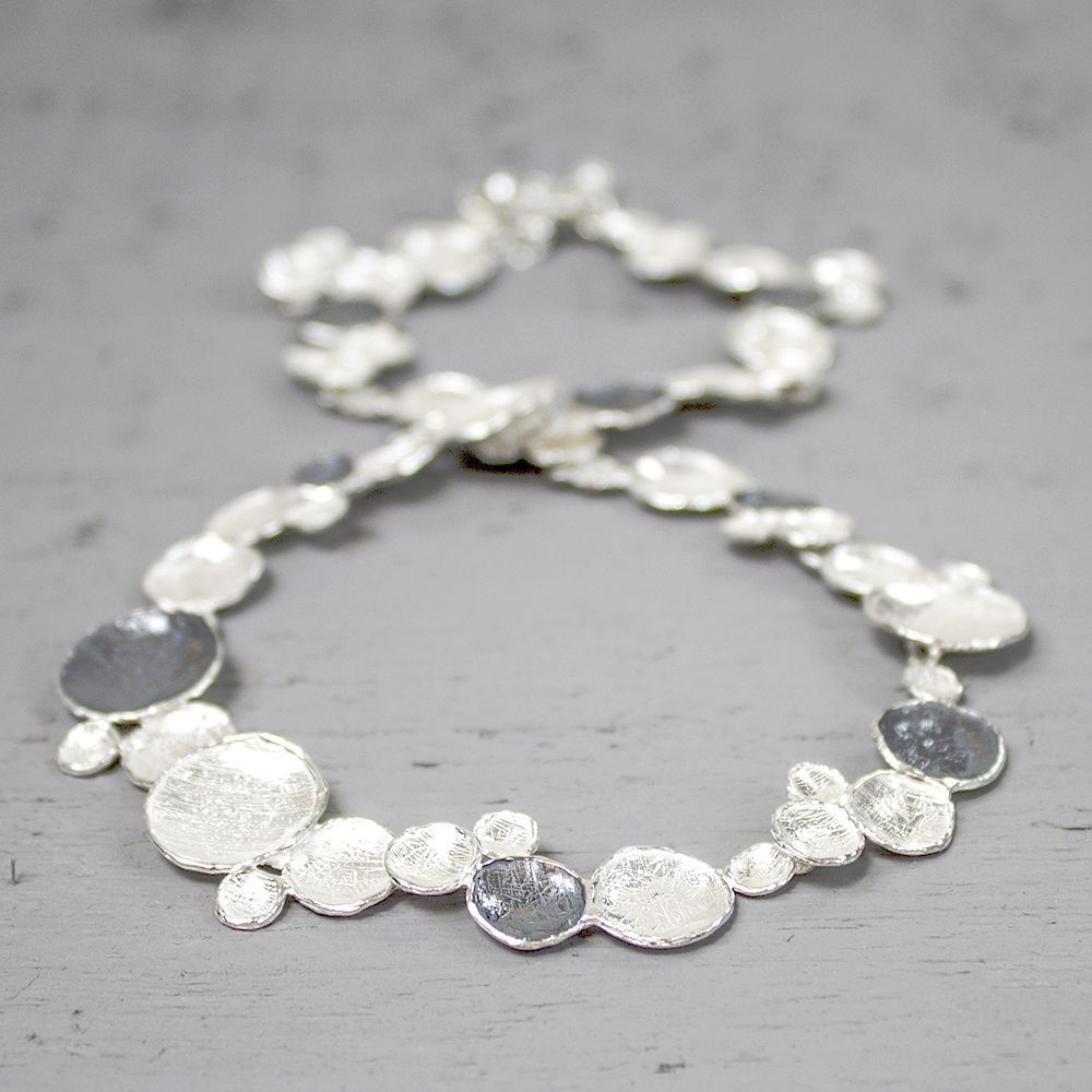 Zilveren ketting met onregelmatig gevormde, gestructureerde hangers op een grijze ondergrond.