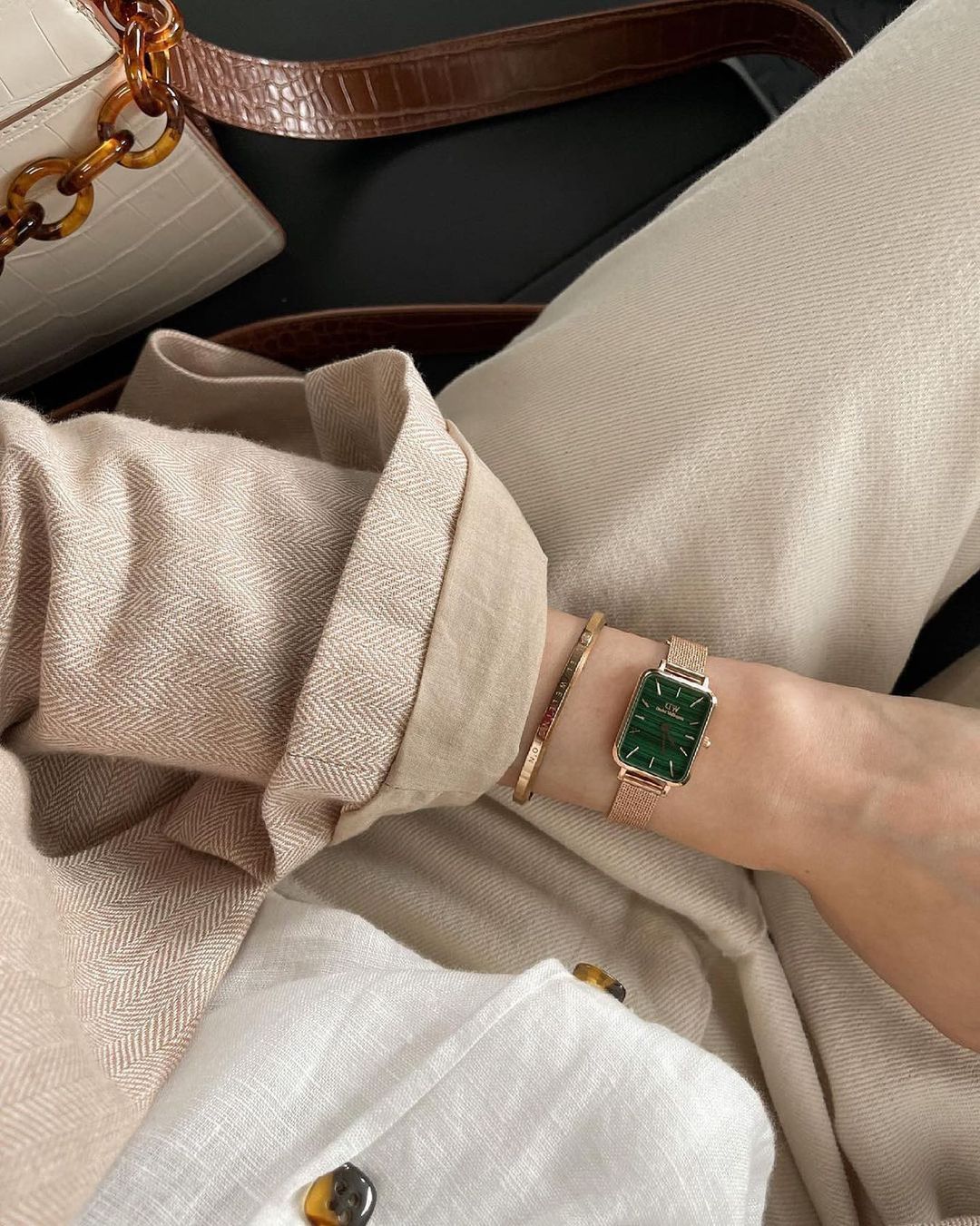 Close-up van de pols van een persoon met een groen horloge en een gouden armband. Beige trui en broek zichtbaar.