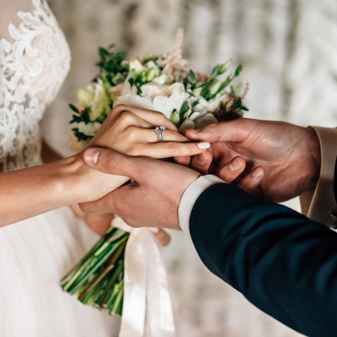 De handen van een bruidspaar dat ringen uitwisselt tijdens een huwelijksceremonie, met een boeket op de achtergrond.