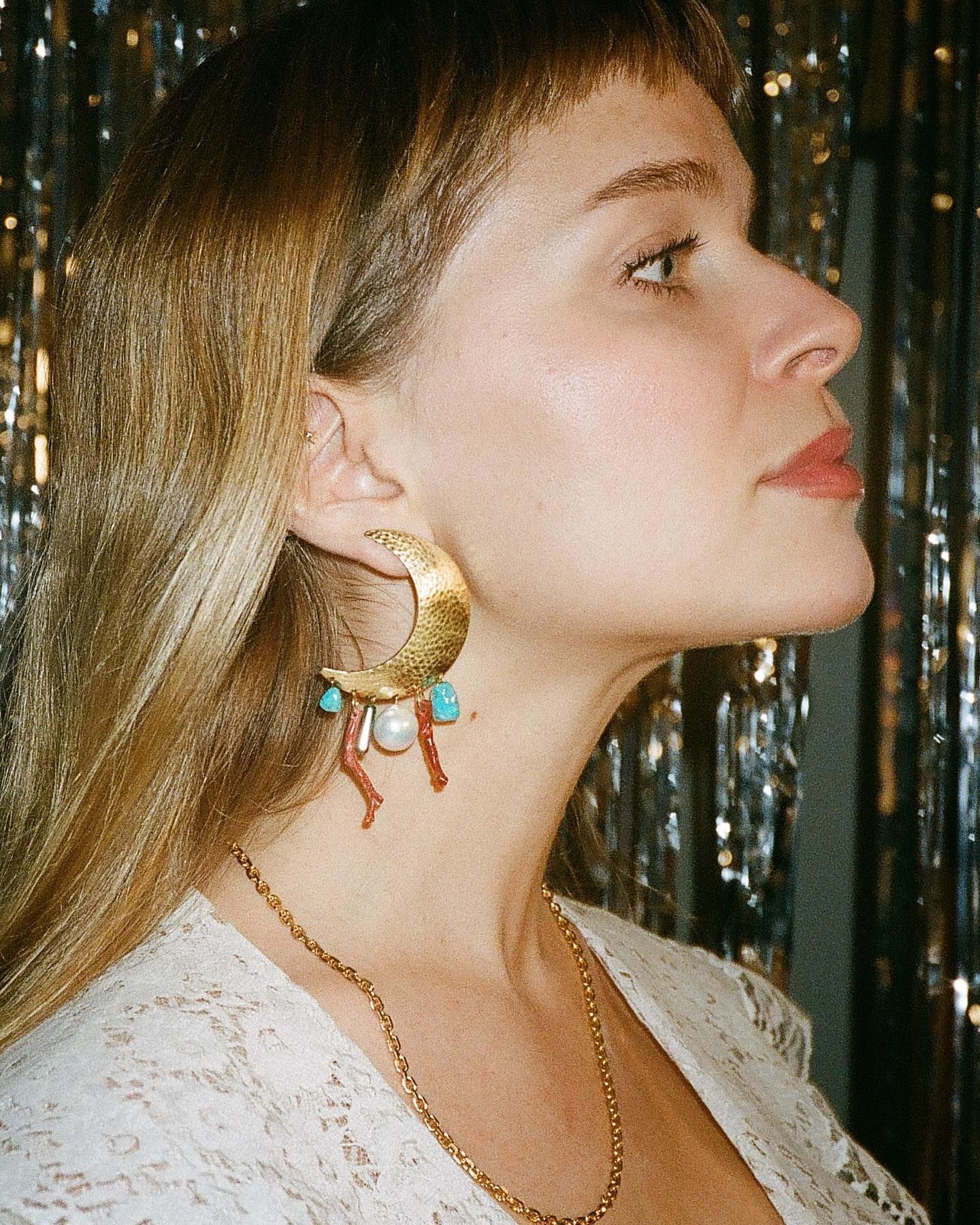 Vrouw in profiel met een halvemaanvormige oorbel met turquoise accenten en een gouden ketting.