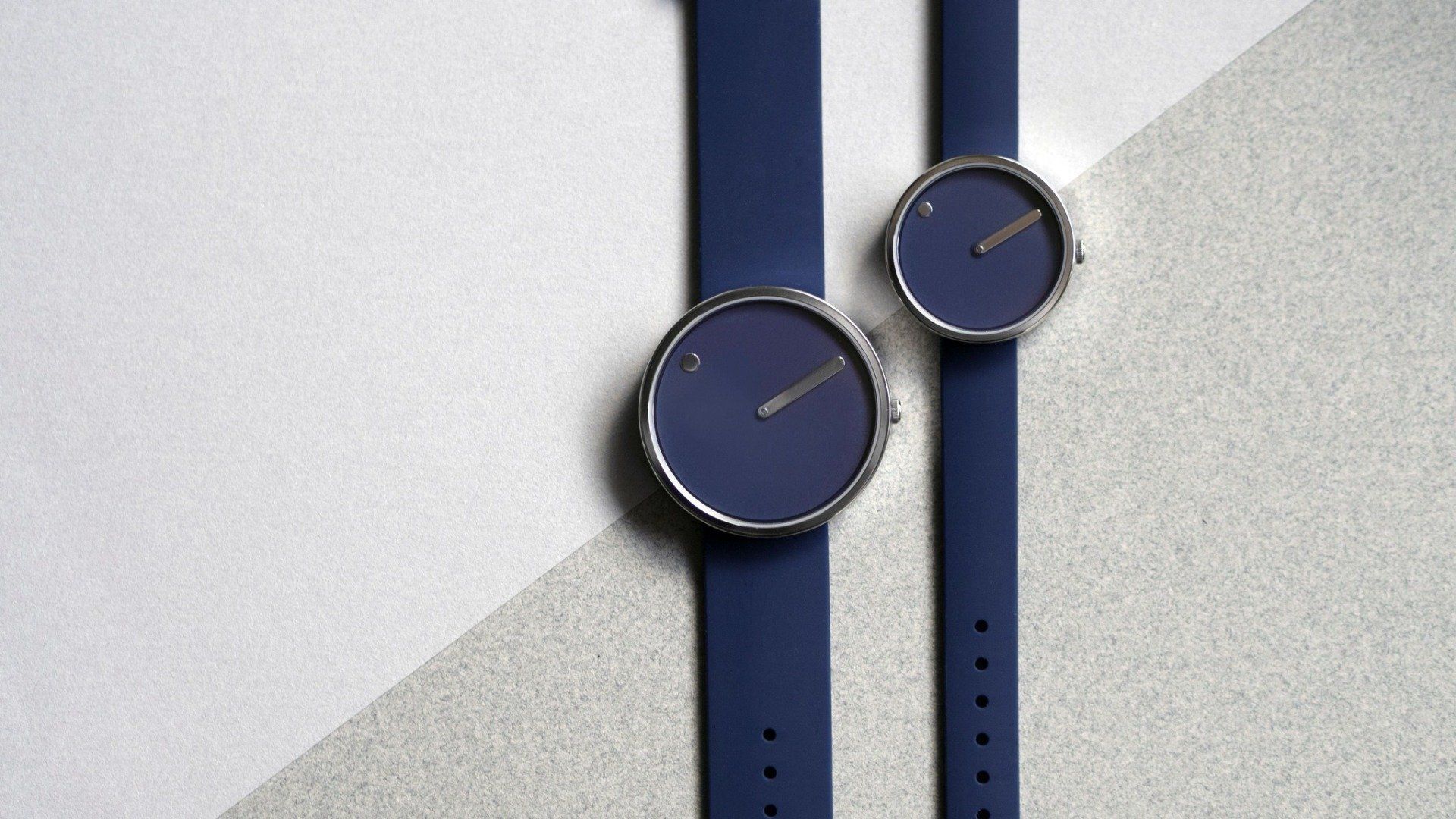 Twee minimalistische blauwe horloges met zilveren randen en banden, op een tweekleurige wit-grijze achtergrond.
