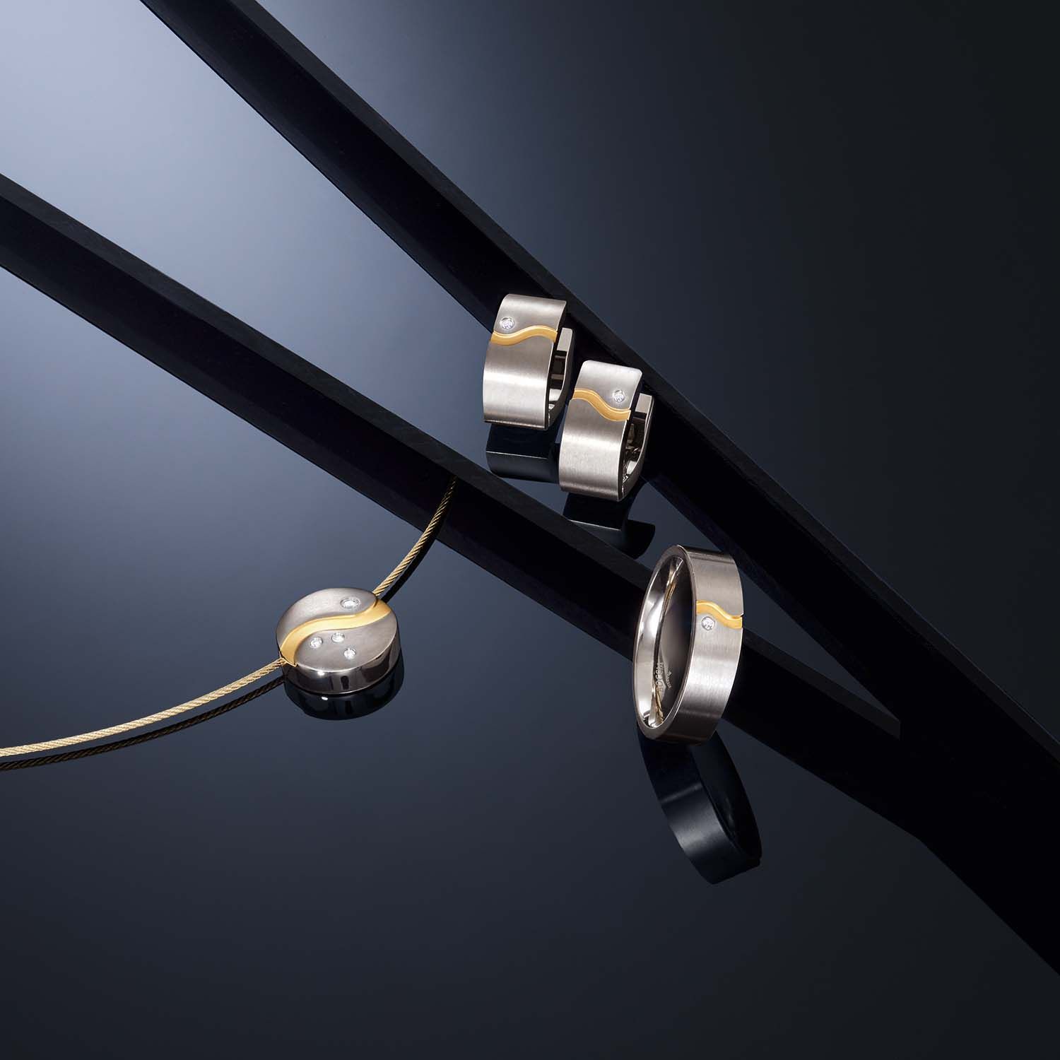 Sieradenset: zilveren ringen en een hanger met gouden accenten op een reflecterend zwart oppervlak.
