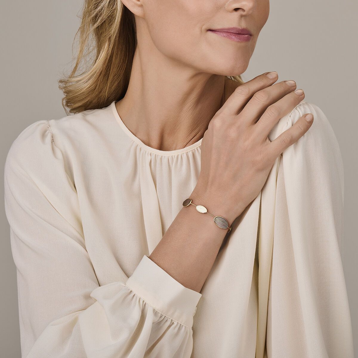 Vrouw draagt een armband, hand op haar schouder, crèmekleurige blouse.