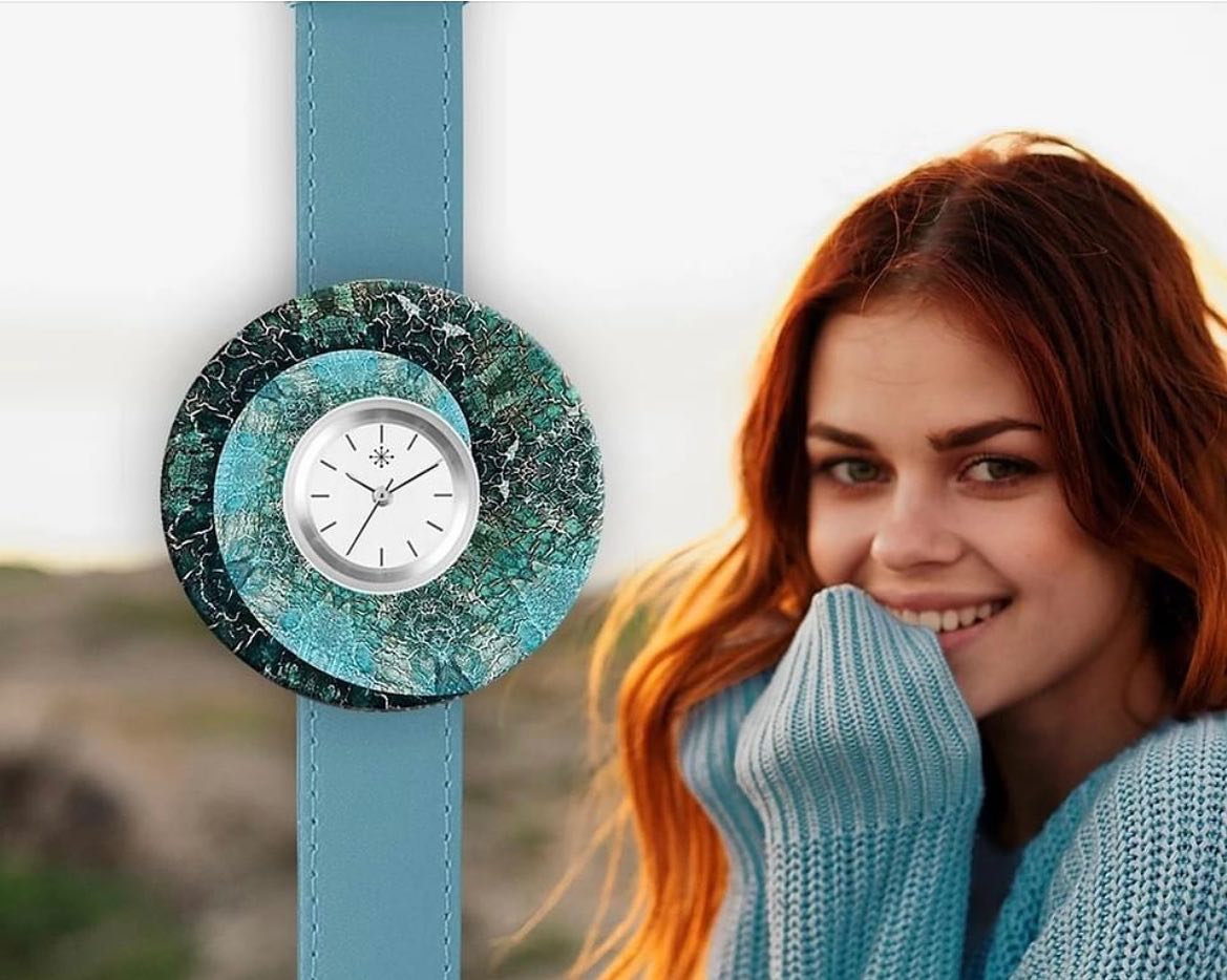 Een vrouw draagt ​​een blauwgroen horloge met een gelaagde, getextureerde wijzerplaat, glimlachend. Ze draagt ​​een blauwe trui.