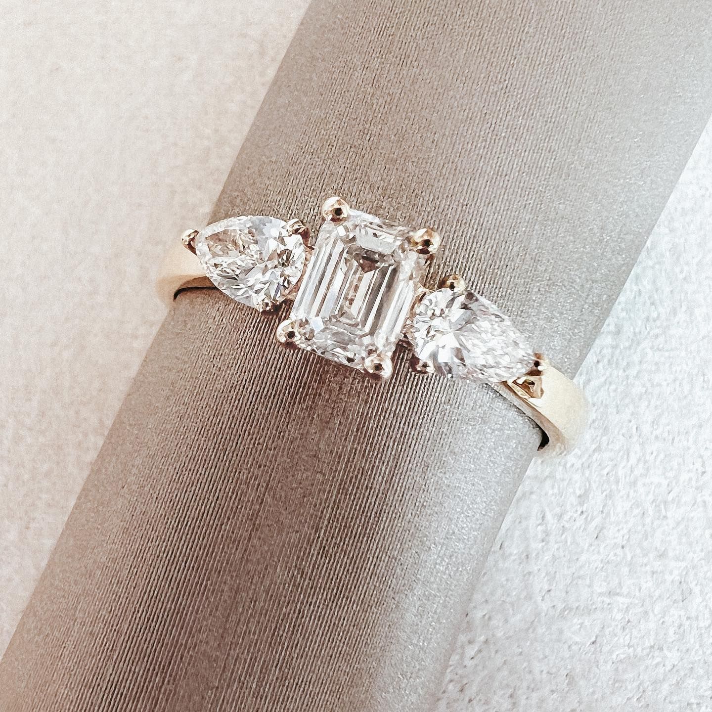 Gouden ring met een smaragdgeslepen diamant in het midden en peervormige zijstenen op een beige stoffen rol.