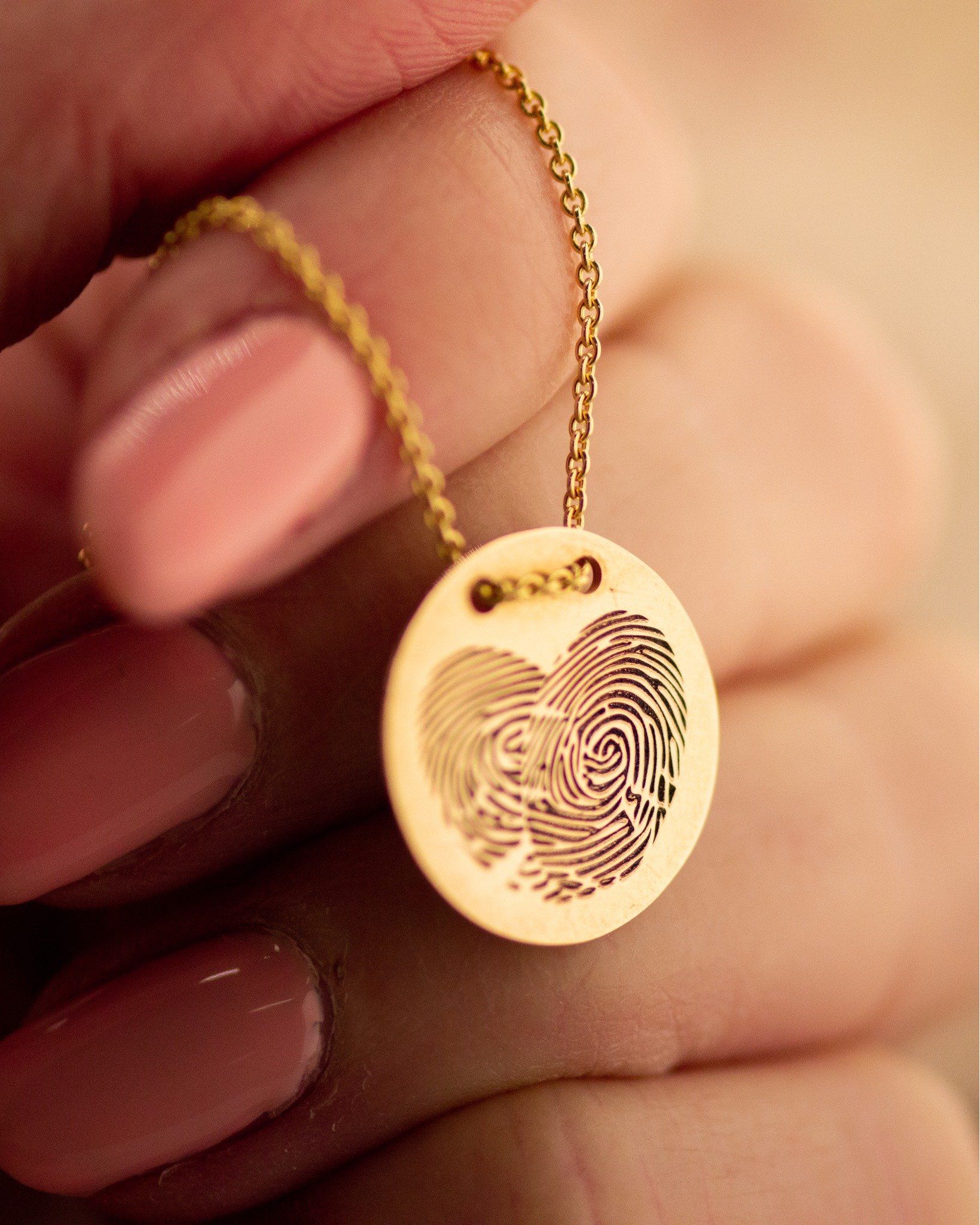 Gouden hanger met vingerafdrukgravure, vastgehouden door een hand met roze nagels.