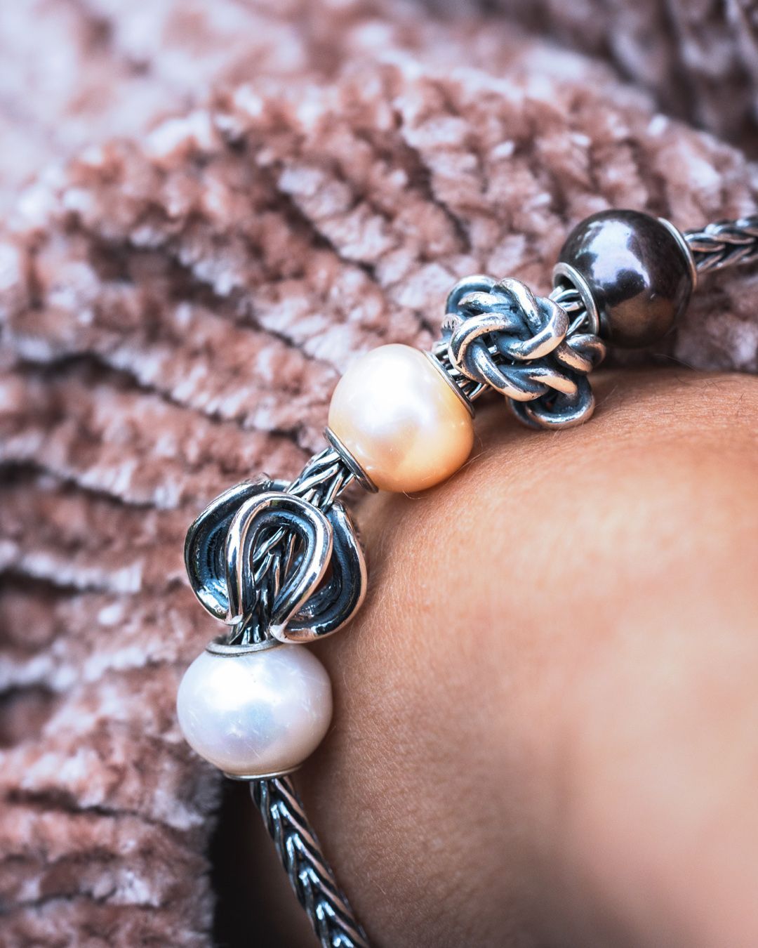 Zilveren armband met parels en knoopvormige bedels om de pols, op een zachte bruine achtergrond.