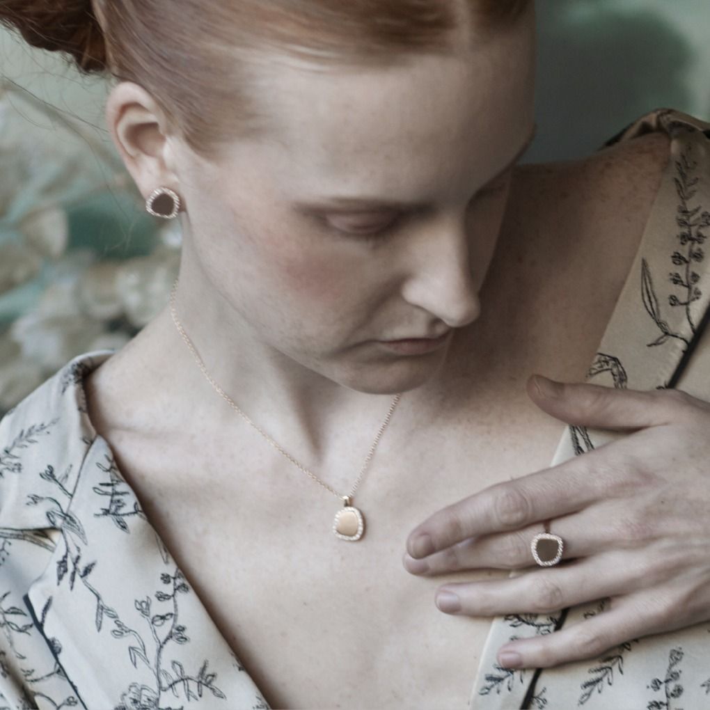 Vrouw met sieraden, naar beneden kijkend. Gouden ketting, oorbellen en ring zichtbaar. Bloemenachtergrond.