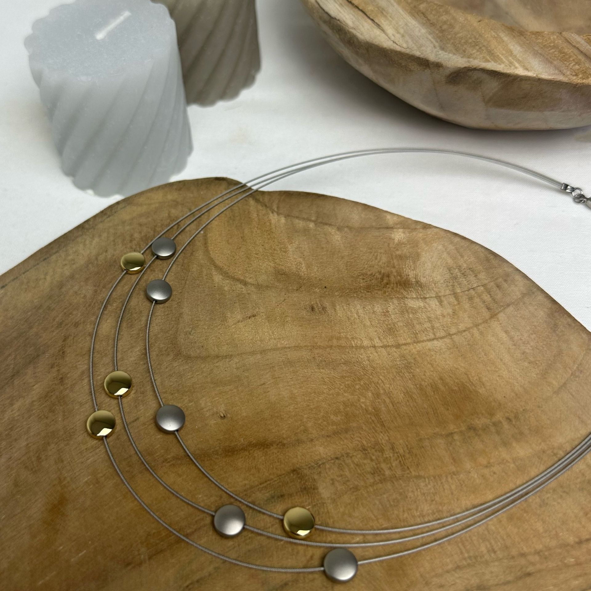 Drie-rijige zilveren ketting met gouden en zilveren schijfaccenten op een houten oppervlak, naast kaarsen.