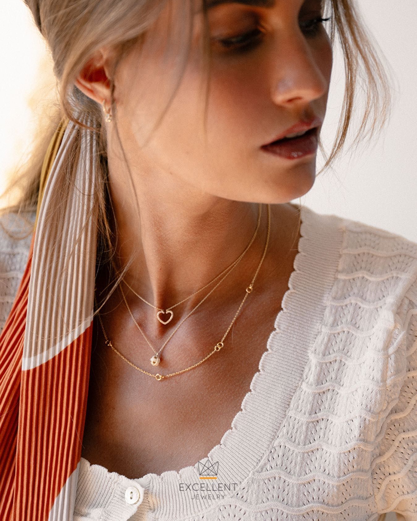 Vrouw draagt ​​drie gouden kettingen en een hartvormige hanger. Crèmekleurige trui en sjaal.