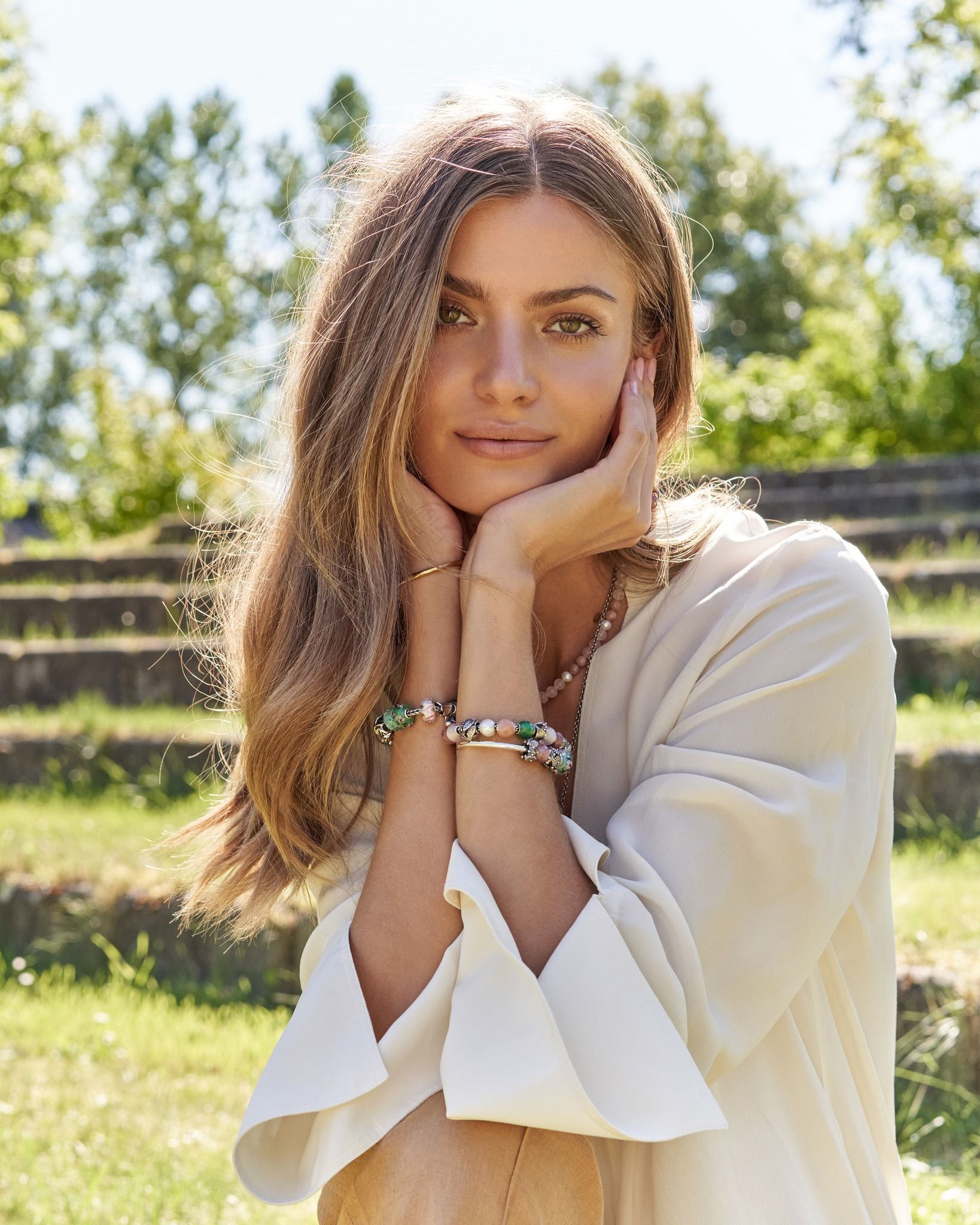 Vrouw met lang golvend haar, kin in haar handen, armbanden om en een wit topje, buiten.