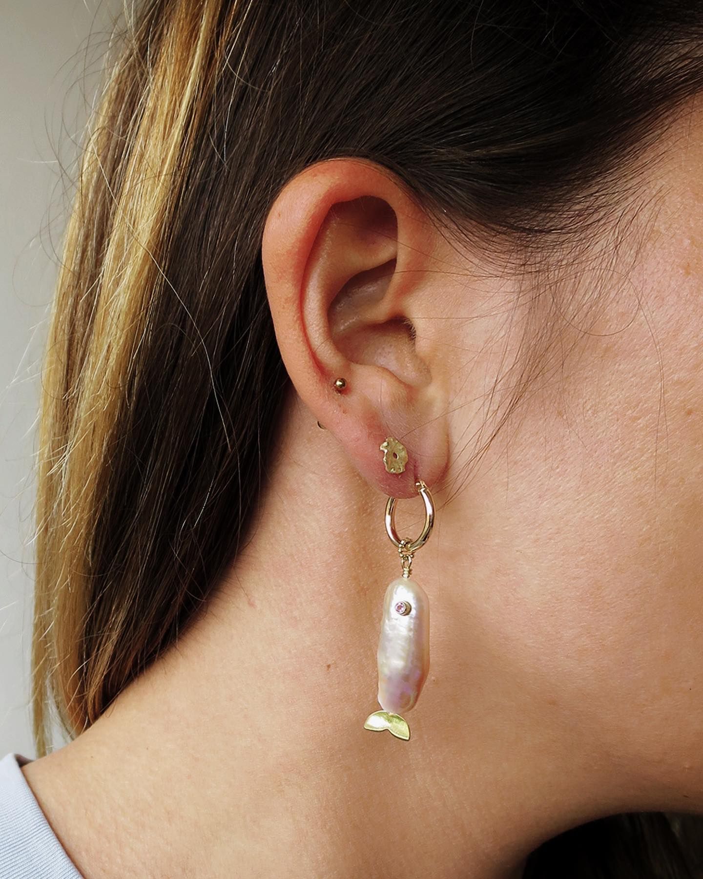 Close-up van het oor van een persoon met een gouden oorbel met een lichtgekleurde parel en een klein gouden bloemenbedeltje.