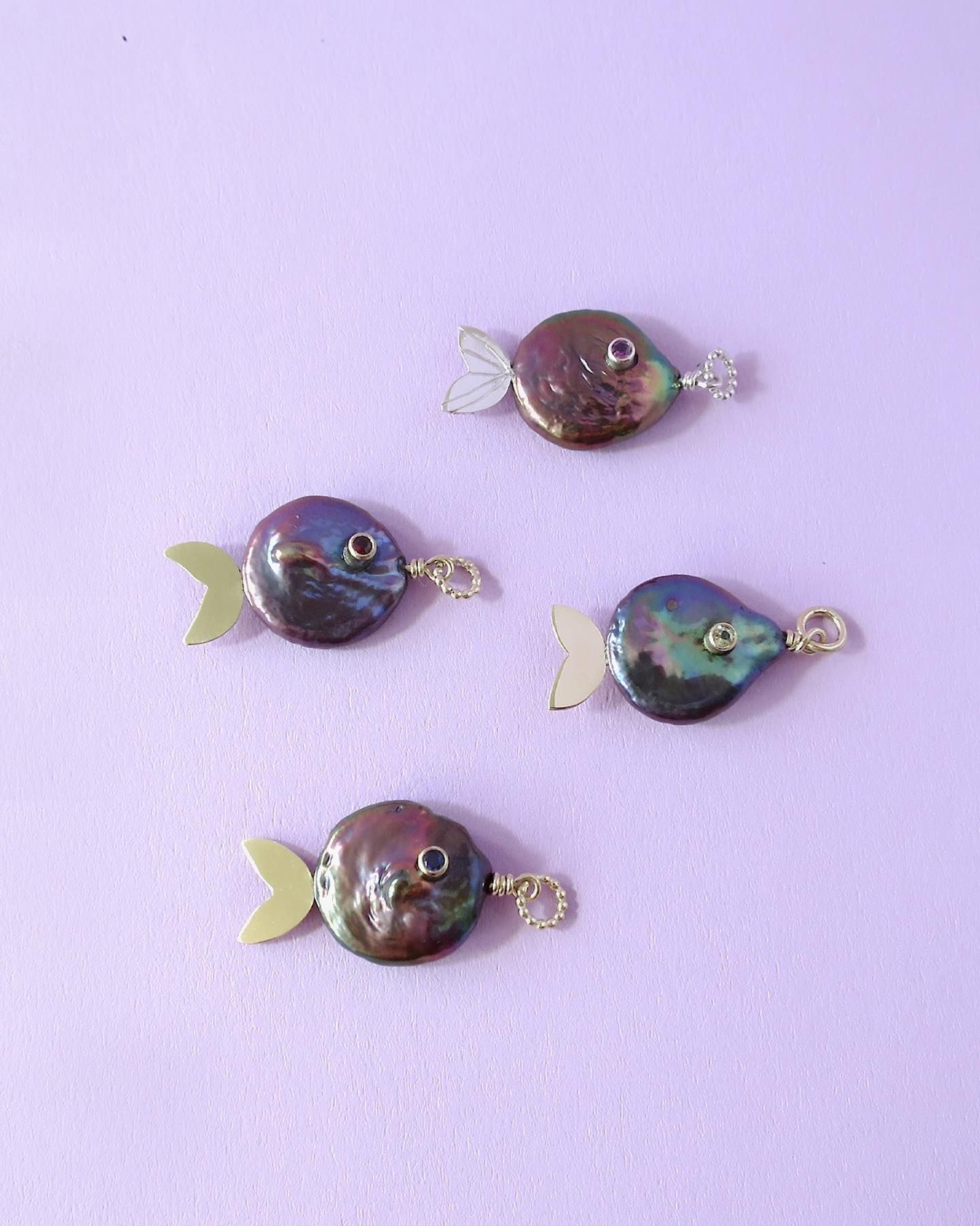 Vier visvormige bedels, gemaakt van donkere iriserende parels met gouden accenten, tegen een lavendelkleurige achtergrond.