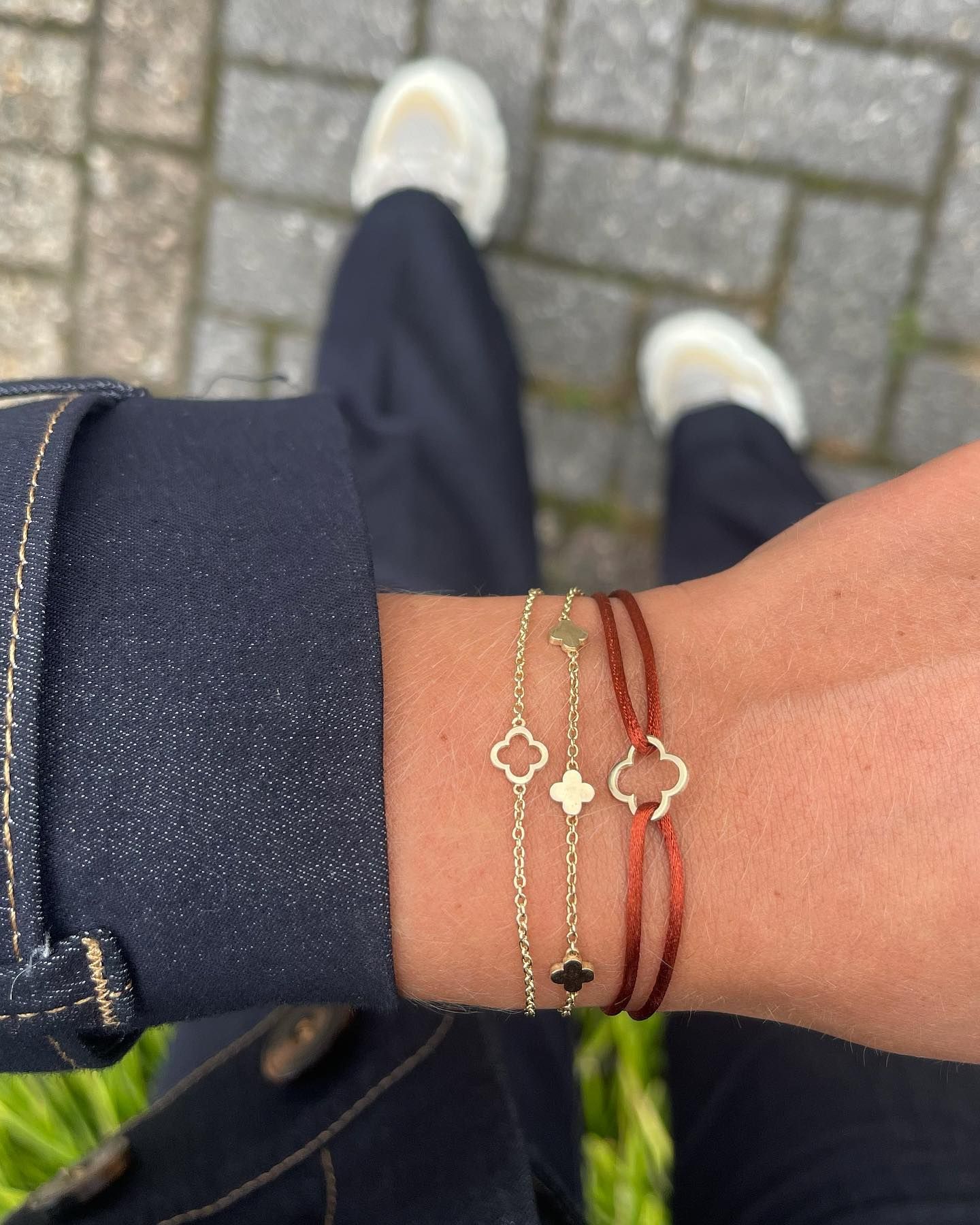 Drie gouden klavertjesarmbanden om een ​​pols, donkerblauwe spijkerbroek, lichte schoenen, grijze baksteen.