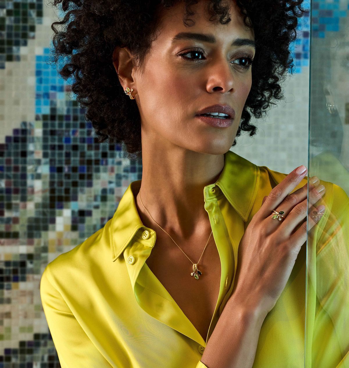 Vrouw draagt gouden sieraden, geel shirt en kijkt opzij, bij een glazen oppervlak.