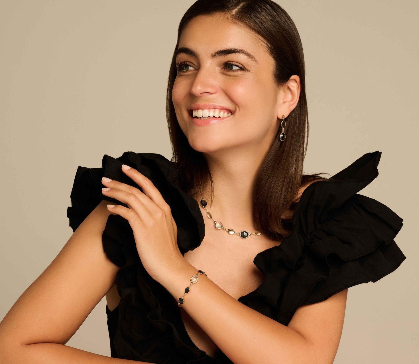 Lachende vrouw in een zwarte jurk, met ketting, armband en oorbellen, tegen een neutrale achtergrond.