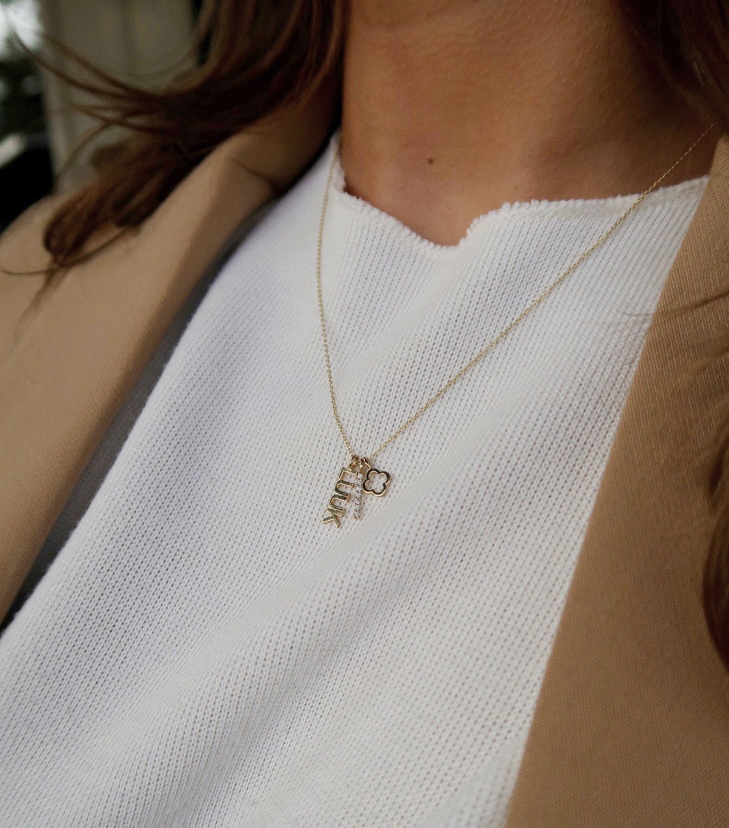Vrouw draagt ​​een gouden ketting met hanger, een witte trui en een bruine jas.