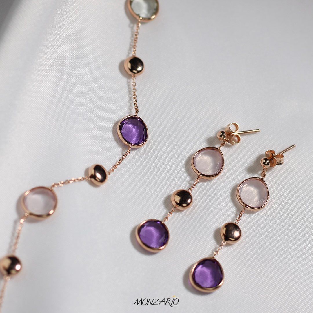 Ketting en oorbellen van roségoud met paarse en lichtgekleurde edelstenen op een witte stoffen achtergrond.