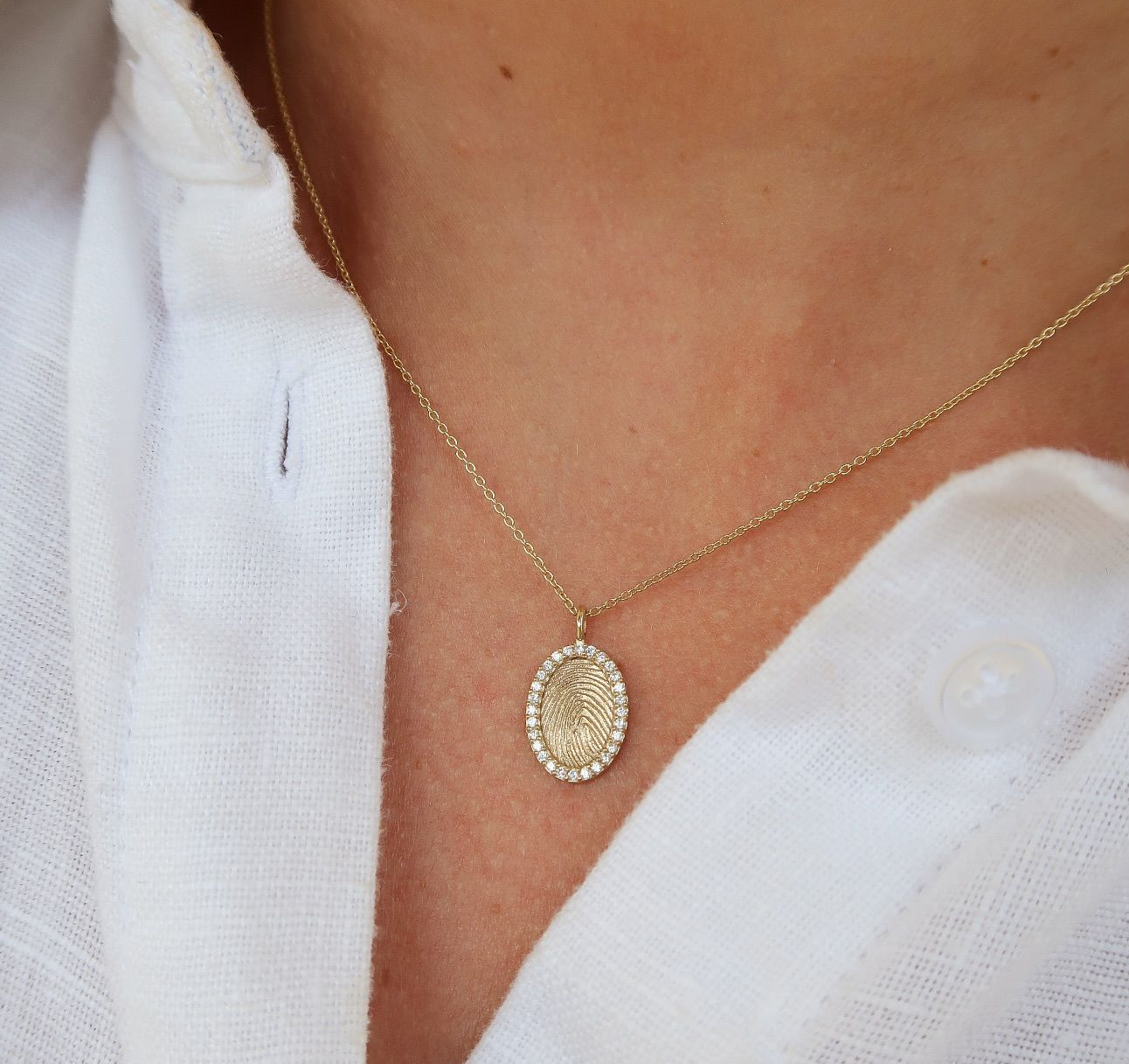 Gouden ketting met ovale hanger bezet met kleine steentjes op een wit shirt.
