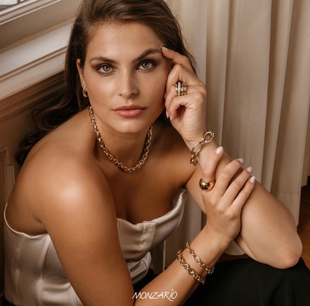 Vrouw in een wit topje met gouden sieraden poseert bij een raam.