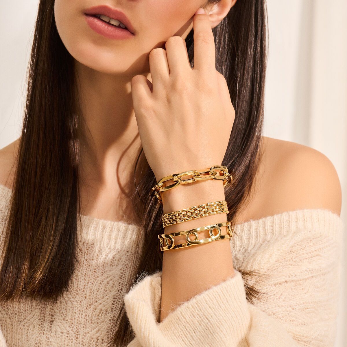 Vrouw draagt meerdere gouden armbanden, houdt haar oor vast en draagt een beige trui met blote schouders.